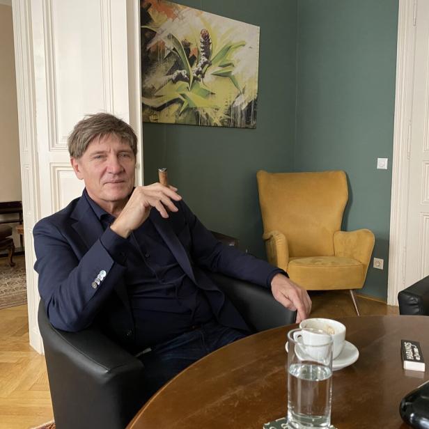 Ein Mann sitzt entspannt in einem Sessel, hält eine Zigarre, vor ihm stehen eine Tasse Kaffee und ein Glas Wasser auf dem Tisch.