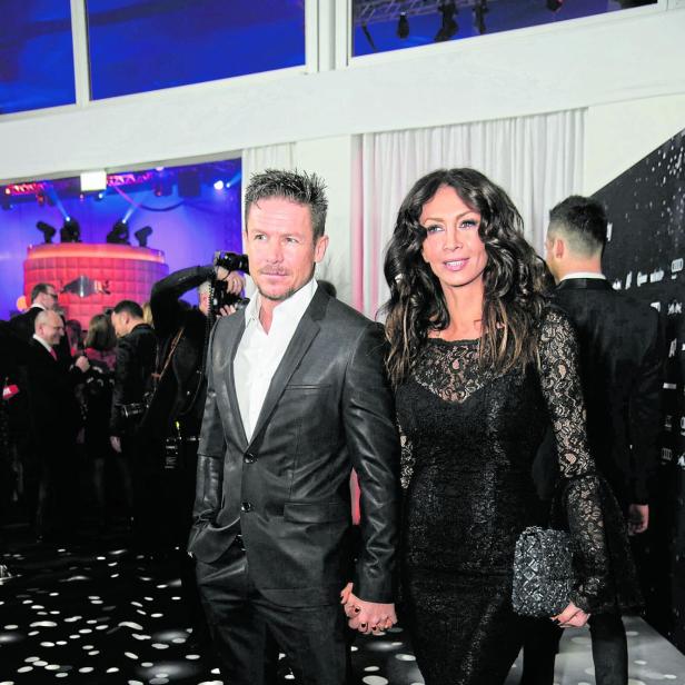 Ein elegant gekleidetes Paar hält Händchen auf einer festlichen Veranstaltung mit vielen Gästen im Hintergrund.