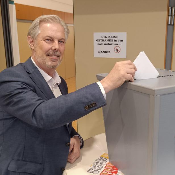 Ein Mann im Anzug wirft einen Wahlzettel in eine graue Wahlurne und lächelt dabei.