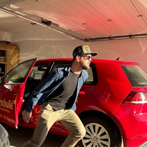 Ein Mann mit Sonnenbrille und Kappe steigt eilig aus einem roten Auto in einer Garage.