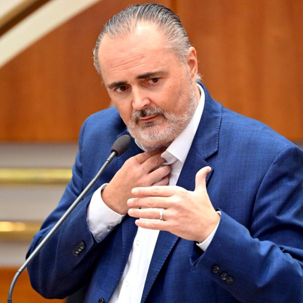 Ein Mann im blauen Anzug spricht gestikulierend an einem Mikrofon in einem Innenraum mit Holzverkleidung.