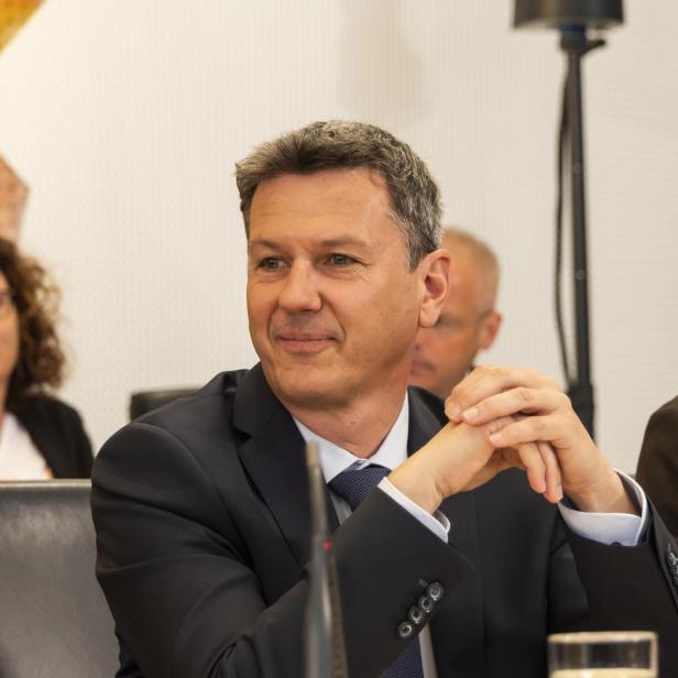Ein Mann im Anzug sitzt an einem Konferenztisch mit Mikrofon und Wasserglas und lächelt leicht.
