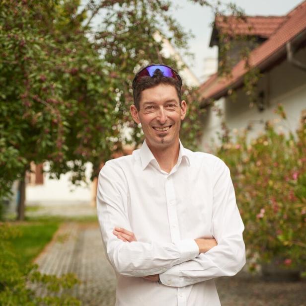 Ein Mann mit weißem Hemd und Sonnenbrille auf dem Kopf steht lächelnd mit verschränkten Armen auf einem gepflasterten Weg im Grünen.