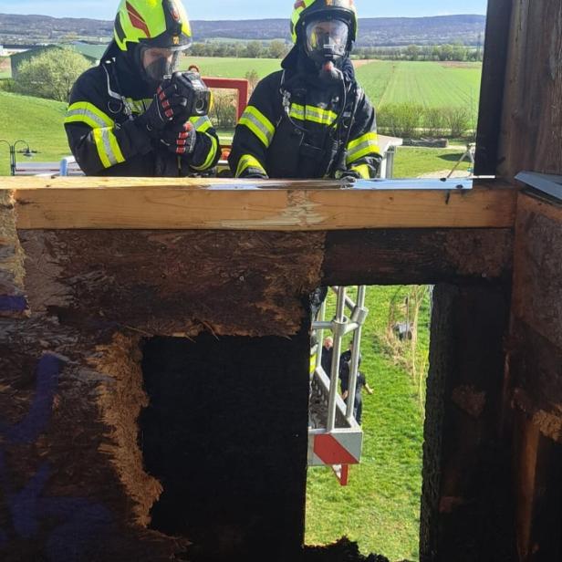 Zwei Feuerwehrleute in Schutzkleidung und Atemschutzmasken stehen an einer verbrannten Holzwand und benutzen eine Wärmebildkamera.