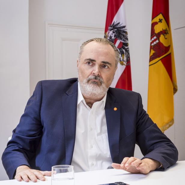 Ein Mann im Anzug sitzt an einem Tisch mit einem Glas Wasser und einem Smartphone, im Hintergrund zwei Fahnen.