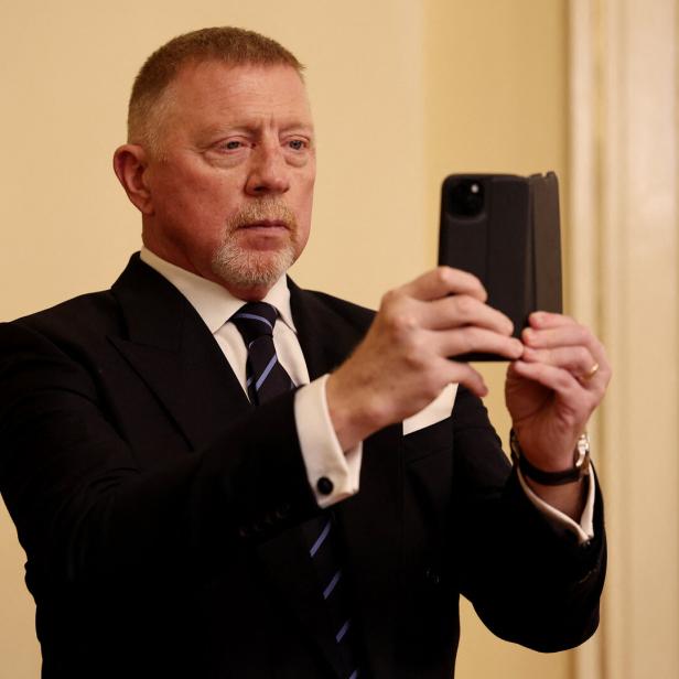 Boris Becker im Anzug hält ein Smartphone hoch.