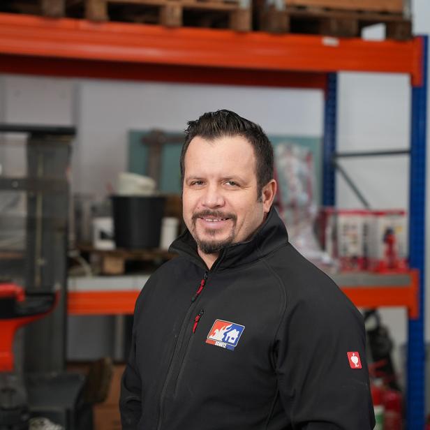 Mann mit schwarzer Jacke steht in einem Lager mit Regalen und Arbeitsmaterialien im Hintergrund.