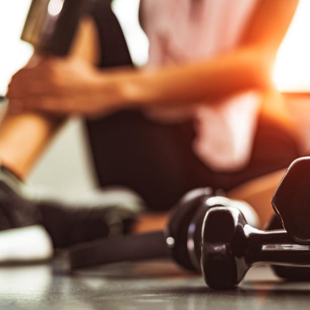 Eine frau sitzt in Sportklamotten auf dem Boden. Vor ihr liegen Hanteln. 