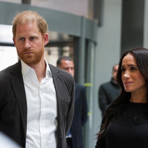 Prinz Harry und Herzogin Meghan, in formeller Kleidung, stehen in einem hellen Innenraum.
