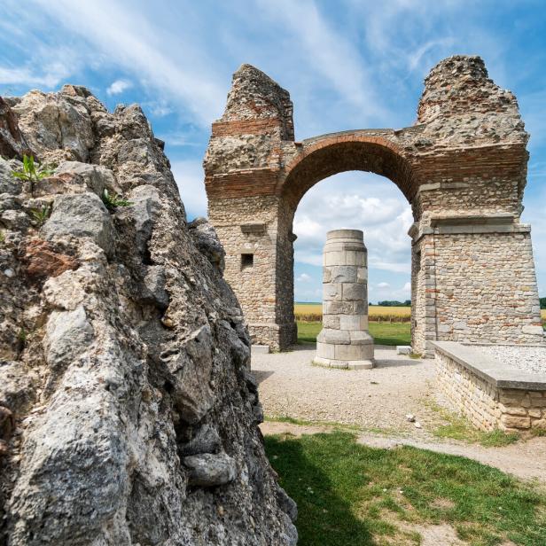 Eine alte Steinruine mit einem Bogen steht vor einem Feld unter blauem Himmel.