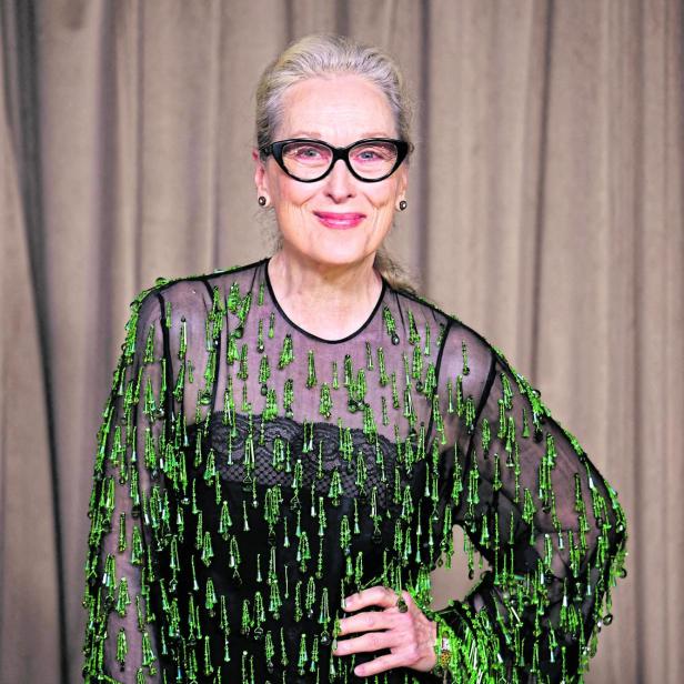 Meryl Streep lächelt mit Brille und grün verziertem, schwarzem Oberteil vor beigem Hintergrund.