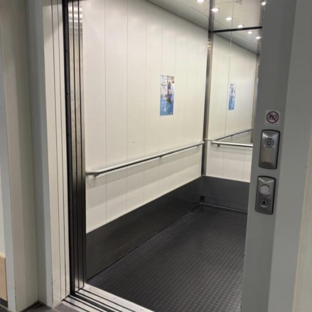 Ein offener Aufzug mit Spiegelwand, Haltegriff und schwarzem Boden ist zu sehen.