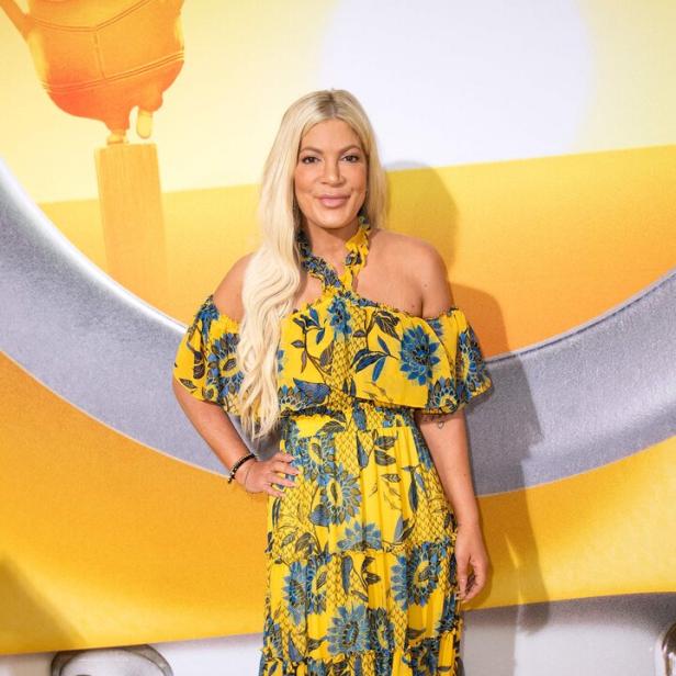 Tori Spelling mit blonden Haaren trägt ein gelbes, geblümtes Kleid vor einem gelben Hintergrund.