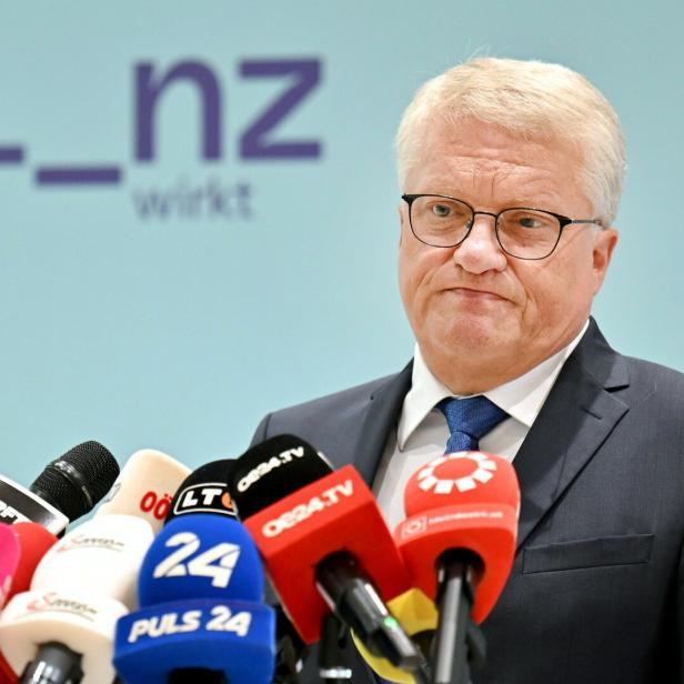 Ein Mann im Anzug steht vor mehreren Mikrofonen und spricht mit ernstem Gesichtsausdruck.