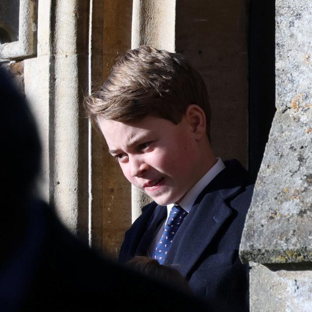 Prinz George mit Anzug und Krawatte steht vor einer steinernen Wand.
