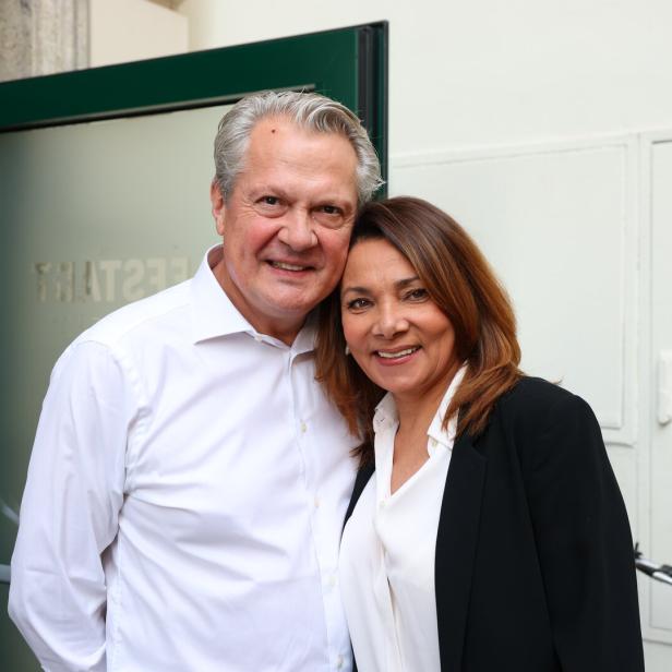 Ein lächelndes Paar, er im weißen Hemd, sie im Blazer, posiert vor einer Tür.