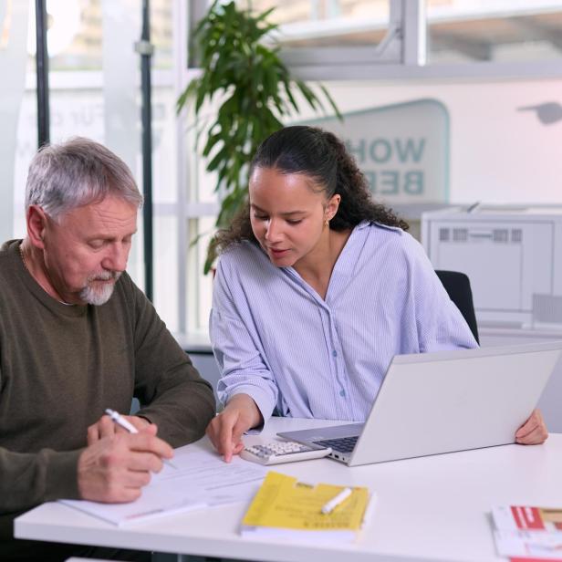 Eine Frau berät einen Mann bei Dokumenten am Schreibtisch mit Laptop und Taschenrechner.