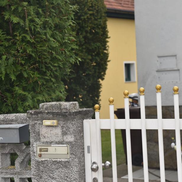 Weiße Gartentür mit goldenen Kugeln, grauer Briefkasten und Klingel an einer Steinmauer, dahinter grüne Hecke und gelbes Haus.
