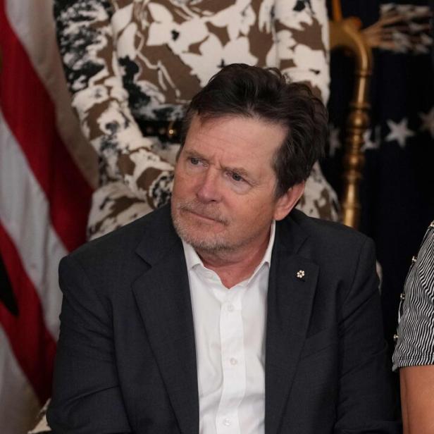 Michael J. Fox sitzt im Anzug, die Hände gefaltet, im Hintergrund Flaggen und Personen.