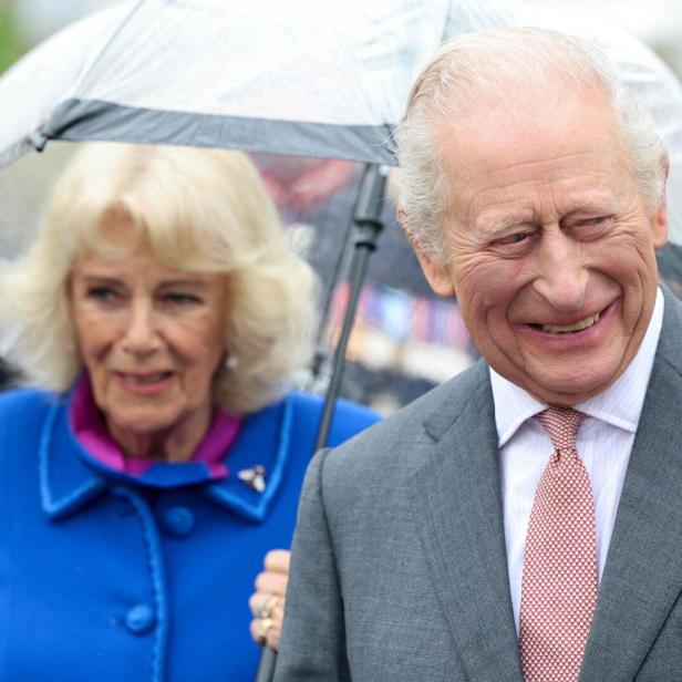 König Charles III. lächelt unter einem Regenschirm, neben ihm Königin Camilla in einem blauen Mantel.
