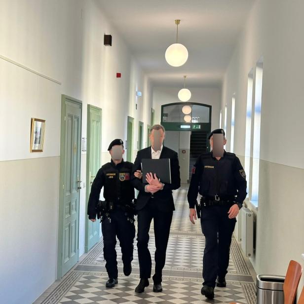 Ein Mann in Anzug wird von zwei Polizisten durch einen hellen Flur begleitet.