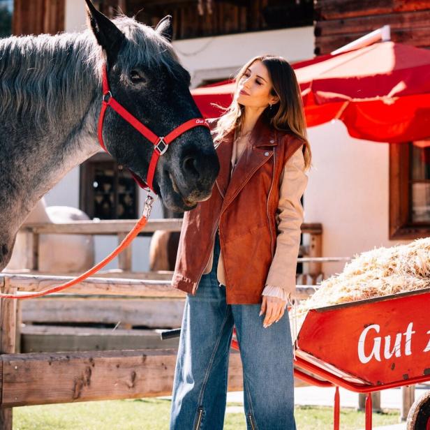 Eine Frau steht neben einem grauen Pferd mit rotem Halfter und einer roten Schubkarre voller Stroh mit der Aufschrift „Gut Aiderbichl“.