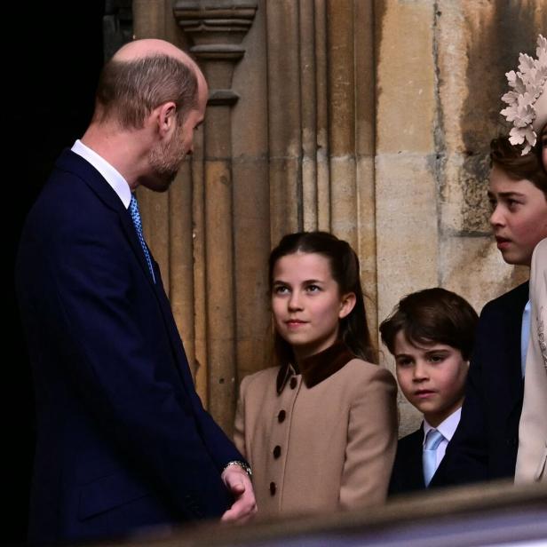 Mehrere elegant gekleidete Personen, darunter Prinz William, Prinzessin Kate und ihre Kinder George, Charlotte und Louis, stehen vor einem steinernen Gebäude und unterhalten sich, eine Frau trägt einen auffälligen Hut.
