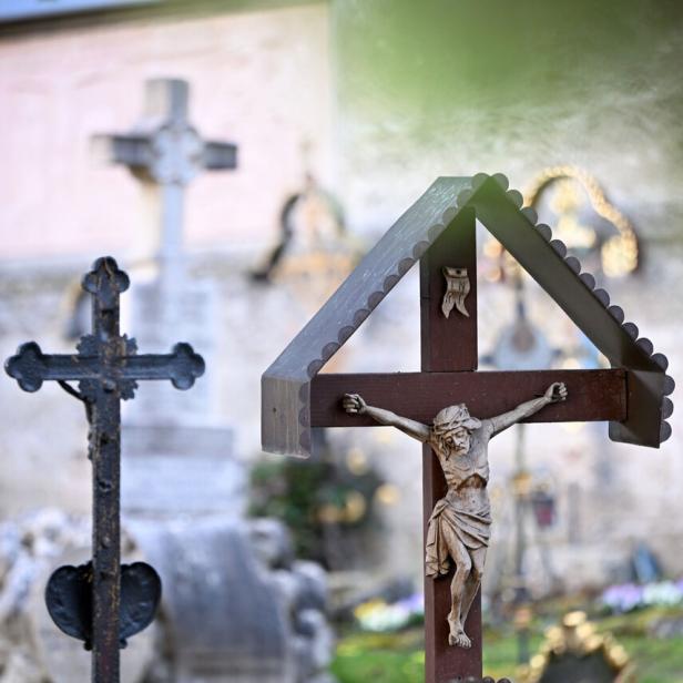 Holzkreuz mit Jesusfigur im Vordergrund, weitere Kreuze und unscharfe Gräber im Hintergrund.