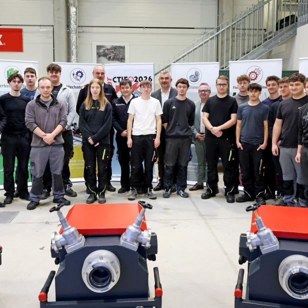 Eine Gruppe von jungen Erwachsenen und mehreren Erwachsenen steht in einer Werkhalle vor technischen Geräten und Informationsplakaten.