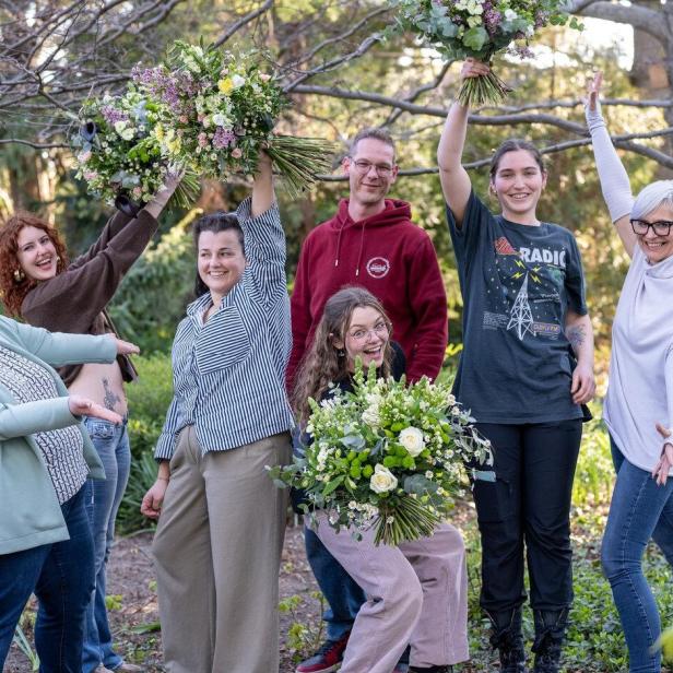 Sieben fröhliche Menschen posieren im Freien, einige halten große Blumensträuße in die Luft.