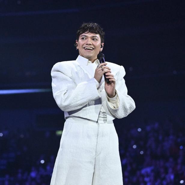 Eine Person in weißem Anzug hält ein Mikrofon und lächelt auf einer Bühne vor großem Publikum.