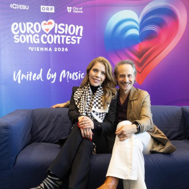 Zwei Personen sitzen vor einem Eurovision Song Contest 2026 Hintergrund auf einem Sofa.