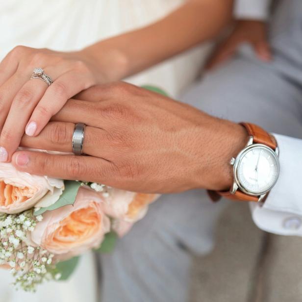 Verschränkte Hände eines Paares mit Eheringen auf einem Brautstrauß mit Rosen.