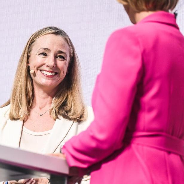Eine lächelnde Frau mit Mikrofon und hellem Blazer steht an einem Tisch, während eine andere Person in Pink daneben steht.