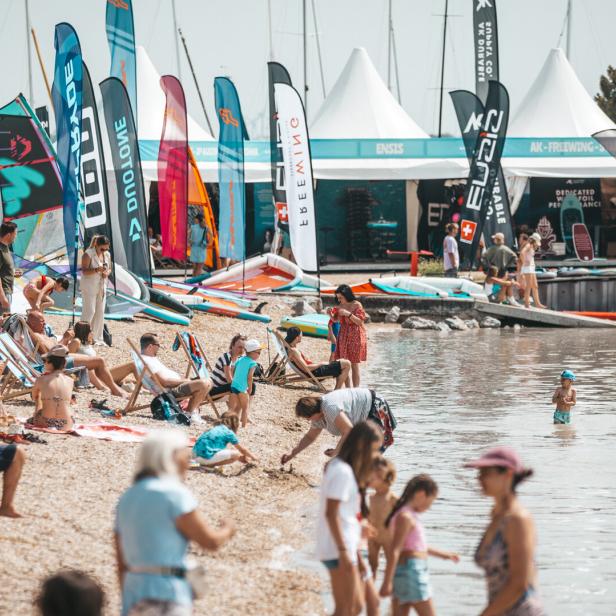 Viele Menschen entspannen am Strand, spielen im Wasser und bunte Fahnen sowie Zelte stehen im Hintergrund.