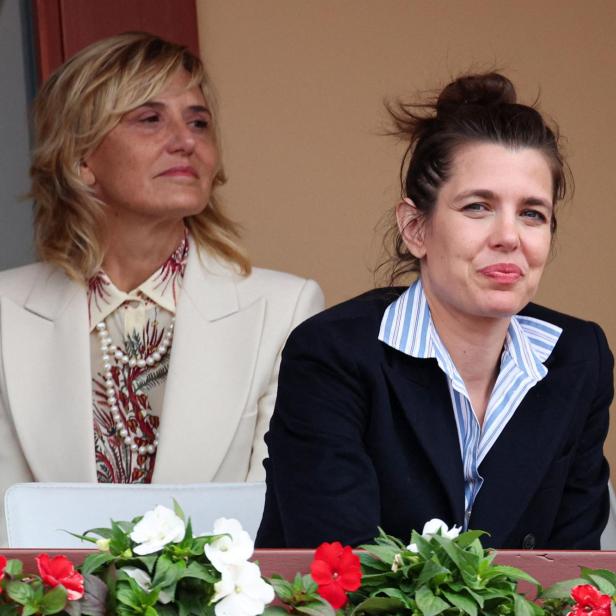 Charlotte Casiraghi sitzt neben einer blonden Frau in einem hellen Blazer vor einem mit Blumen geschmückten Balkongeländer.