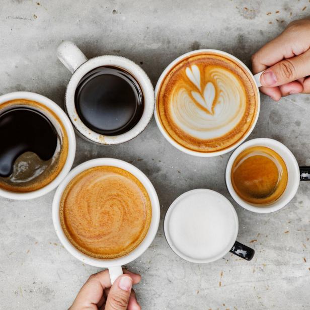 Sechs verschiedene Tassen Kaffee und Milchgetränke stehen auf einer grauen Oberfläche, zwei Hände halten jeweils eine Tasse.