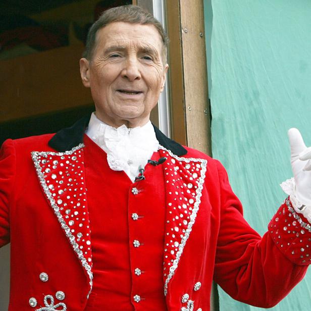 Freddy Quinn trägt einen roten, mit Strass besetzten Frack und weiße Handschuhe und lächelt mit erhobenen Händen.