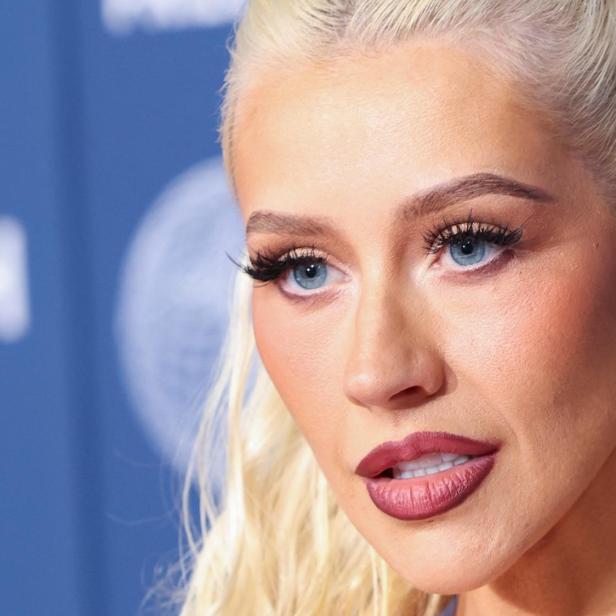 Christina Aguilera mit blonden Haaren, blauem Lidschatten und Ohrringen vor blauem Hintergrund.