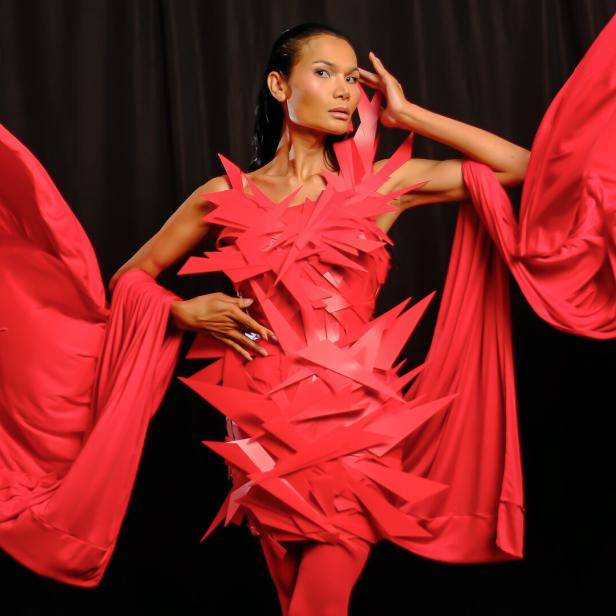 Eine Frau posiert in einem roten Kleid mit zackigen Details und wehenden Stoffbahnen vor schwarzem Hintergrund.