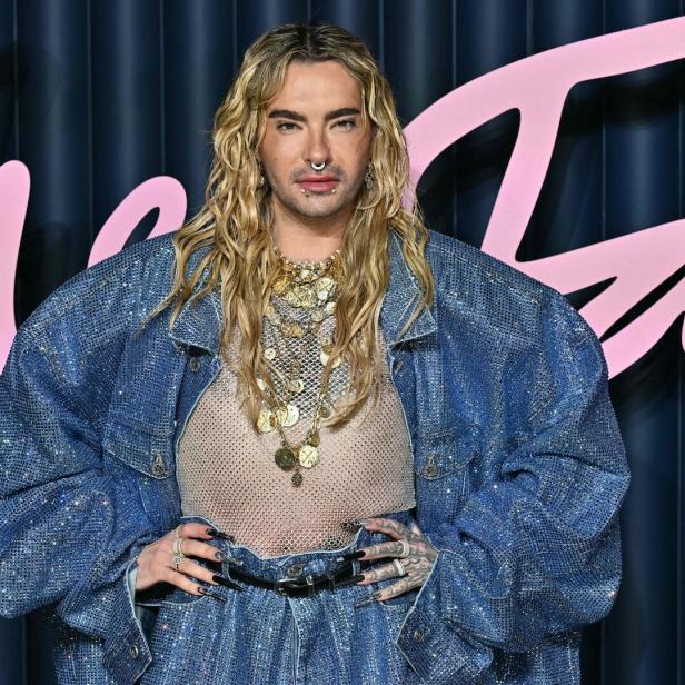 Bill Kaulitz mit blonden, gewellten Haaren, Nasenring und vielen Halsketten trägt eine glitzernde Jeansjacke.