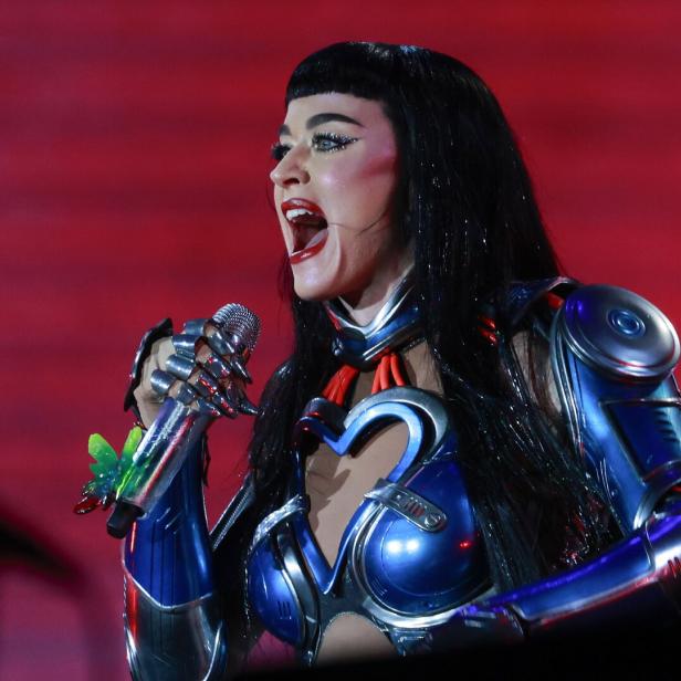 Katy Perry mit schwarzem Haar und futuristischem Kostüm singt vor rotem Hintergrund.