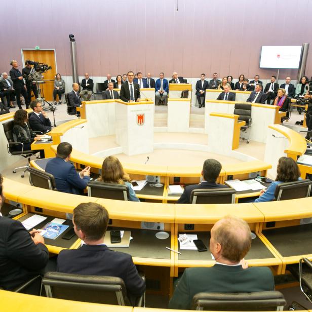 Mehrere Personen sitzen in einem modernen Plenarsaal, während eine Person am Rednerpult spricht.