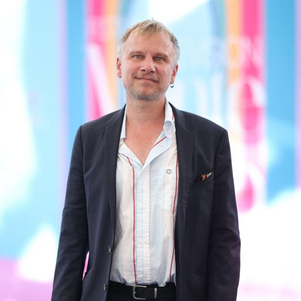 Robert Stadlober mit Sakko und gestreiftem Hemd steht vor einem farbenfrohen Hintergrund.