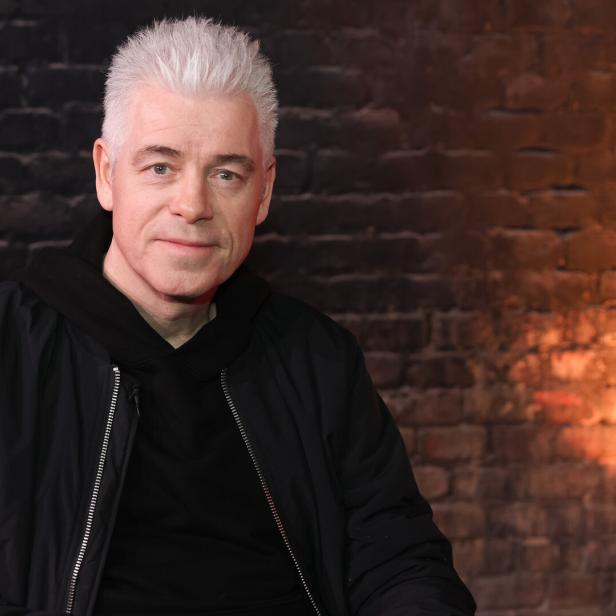 Michael Mittermeier trägt eine schwarze Jacke und sitzt vor einer Backsteinwand mit warmem Licht.