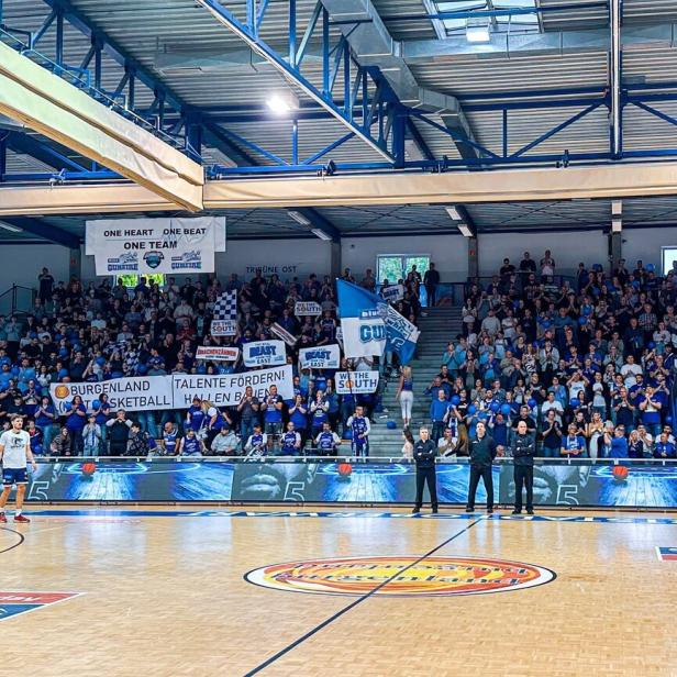 Viele Fans in blauer Kleidung und mit Bannern feuern ein Basketballteam in einer vollen Sporthalle an.