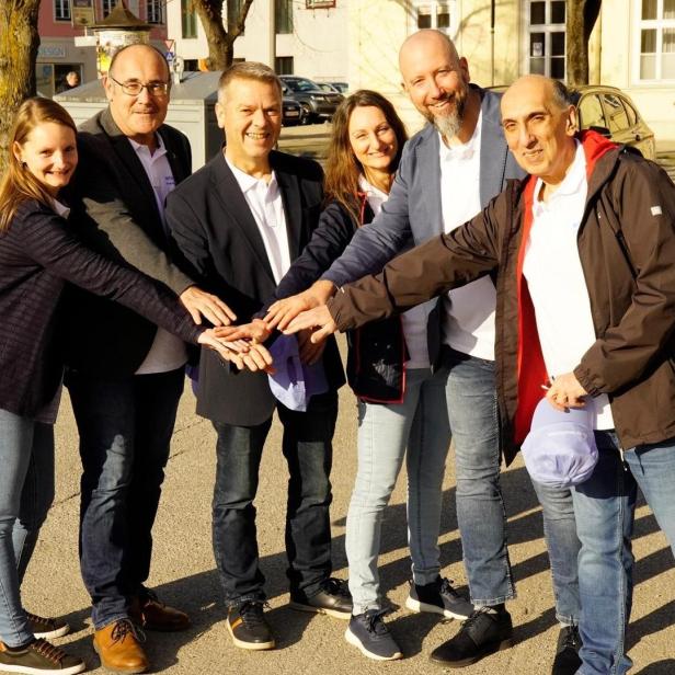 Sechs Erwachsene stehen im Kreis und legen ihre Hände übereinander, alle lächeln und wirken freundlich.