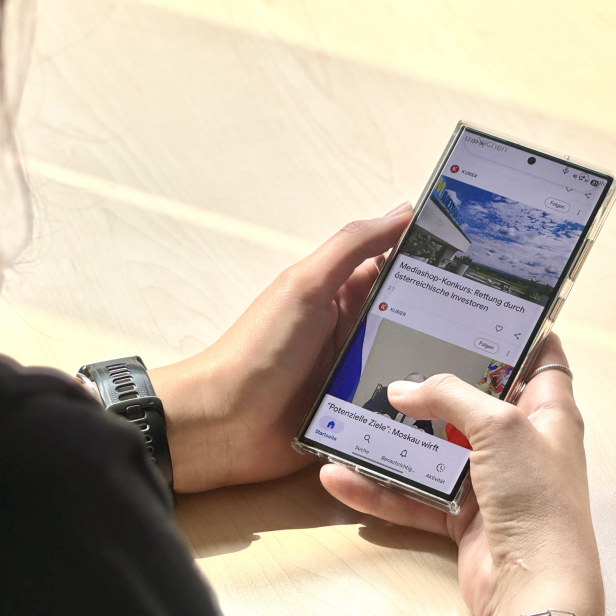 Person hält ein Smartphone und scrollt durch den Google-Discover-Feed mit KURIER-Artikeln.