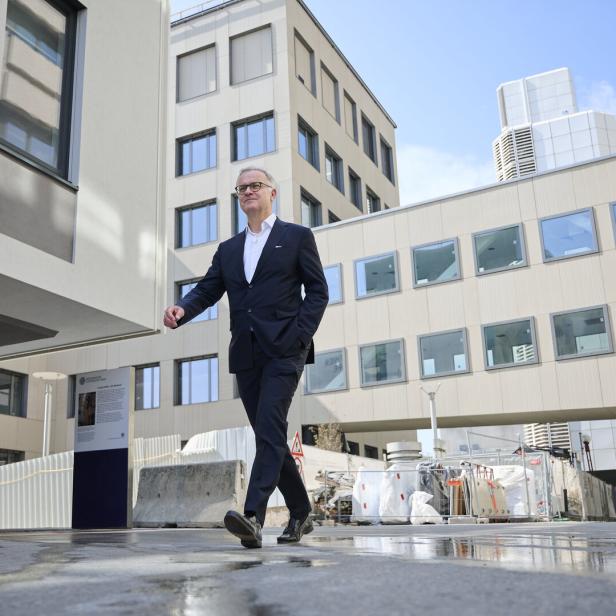 Ein Mann im Anzug geht selbstbewusst durch einen modernen Gebäudekomplex mit Baustellenbereich.