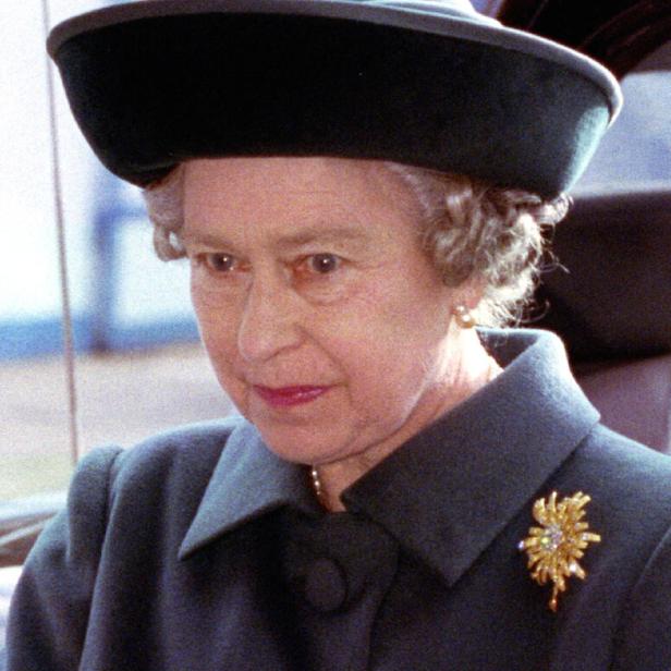 Queen Elizabeth II. mit elegantem dunklen Mantel, großem Hut und goldener Brosche, schaut konzentriert nach vorne.